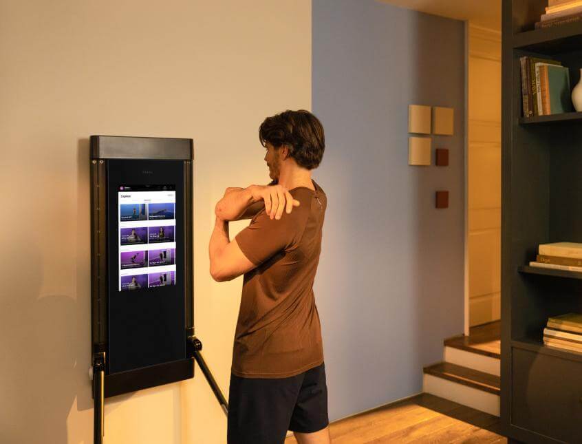 A man stands in his living room. He is in front of his Tonal and stretching—with one arm crossed over his chest. He is looking at the Tonal screen.