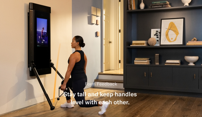 A woman is working out in her living room. She is facing Tonal and is completing an Iso Split Squat.